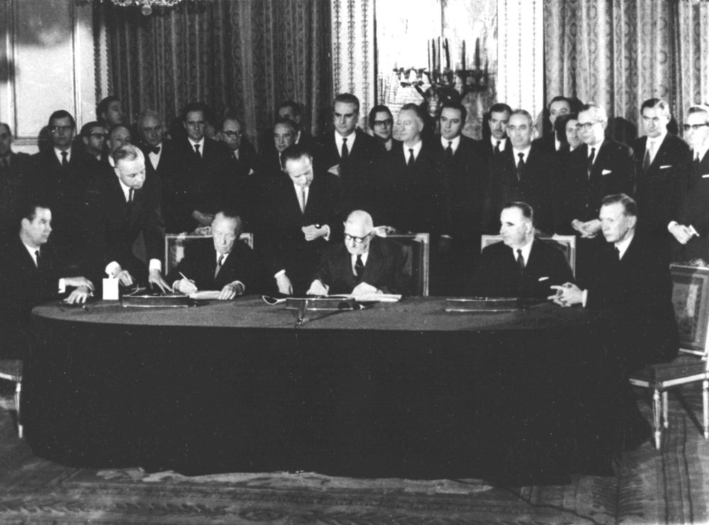 Schwarz-weiß-Fotografie. Im Vordergrund sitzen an einem ovalen Tisch Bundeskanzler Konrad Adenauer und der Staatspräsident Charles de Gaulle, die den deutsch-französischen Freundschaftsvertrag im Pariser Élysée-Palast am 22. Januar 1963 unterzeichnen.
