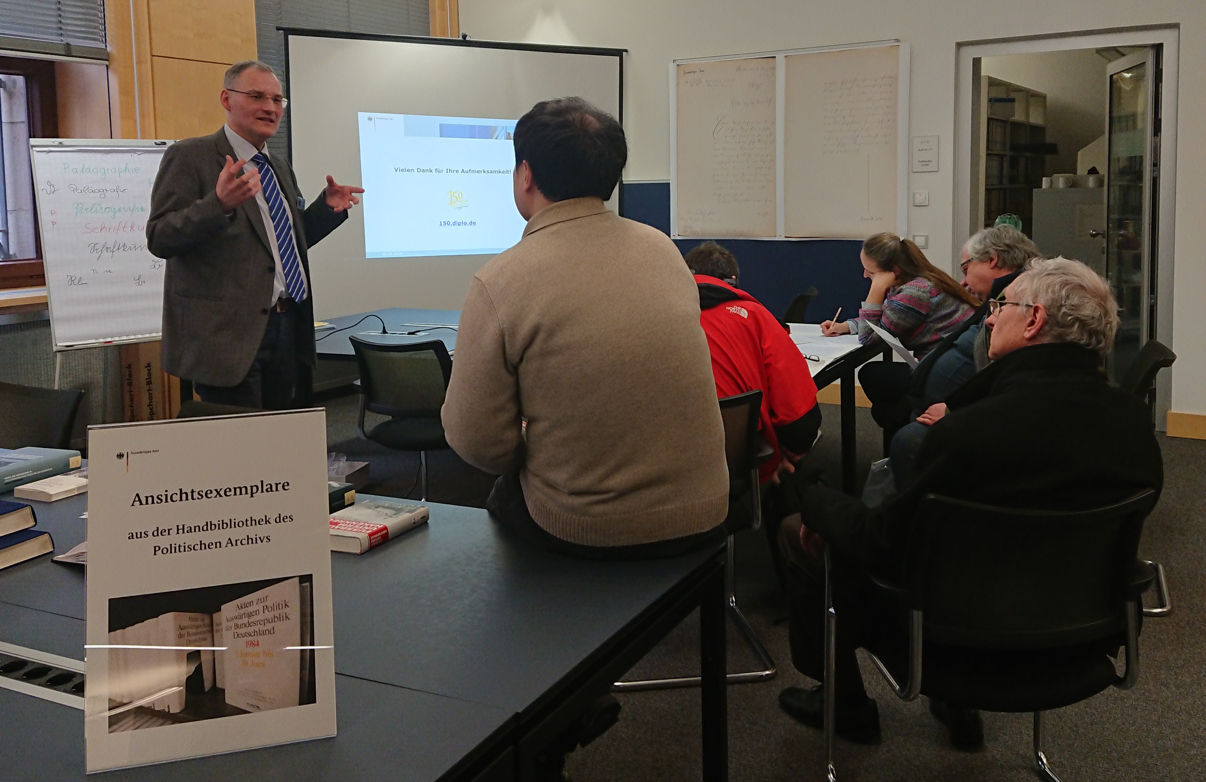 Dr. Simon Heßdörfer erläutert die Entwicklung der Schrift. Die Gäste sitzen am Tisch und schauen auf eine Leinwand mit einer Präsentation.